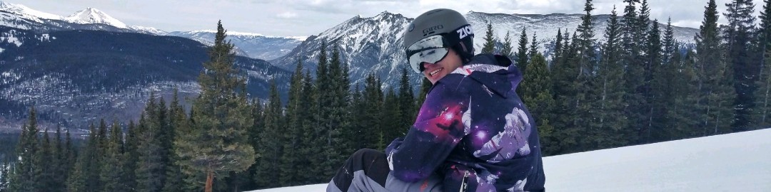 Banner of Leigh sitting on his snowboard, smiling, taking a break, with mountains in the background